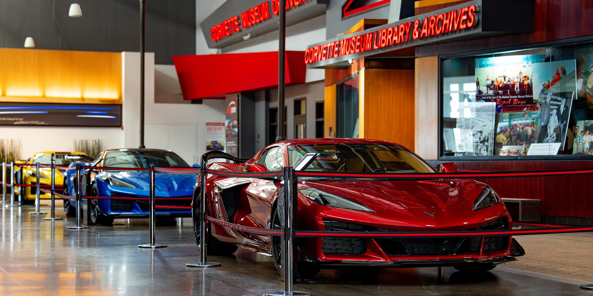 A Red Chevy Corvette Displayed Inside a Showroom Next to Blue Corvette Models Under Bright Lighting.