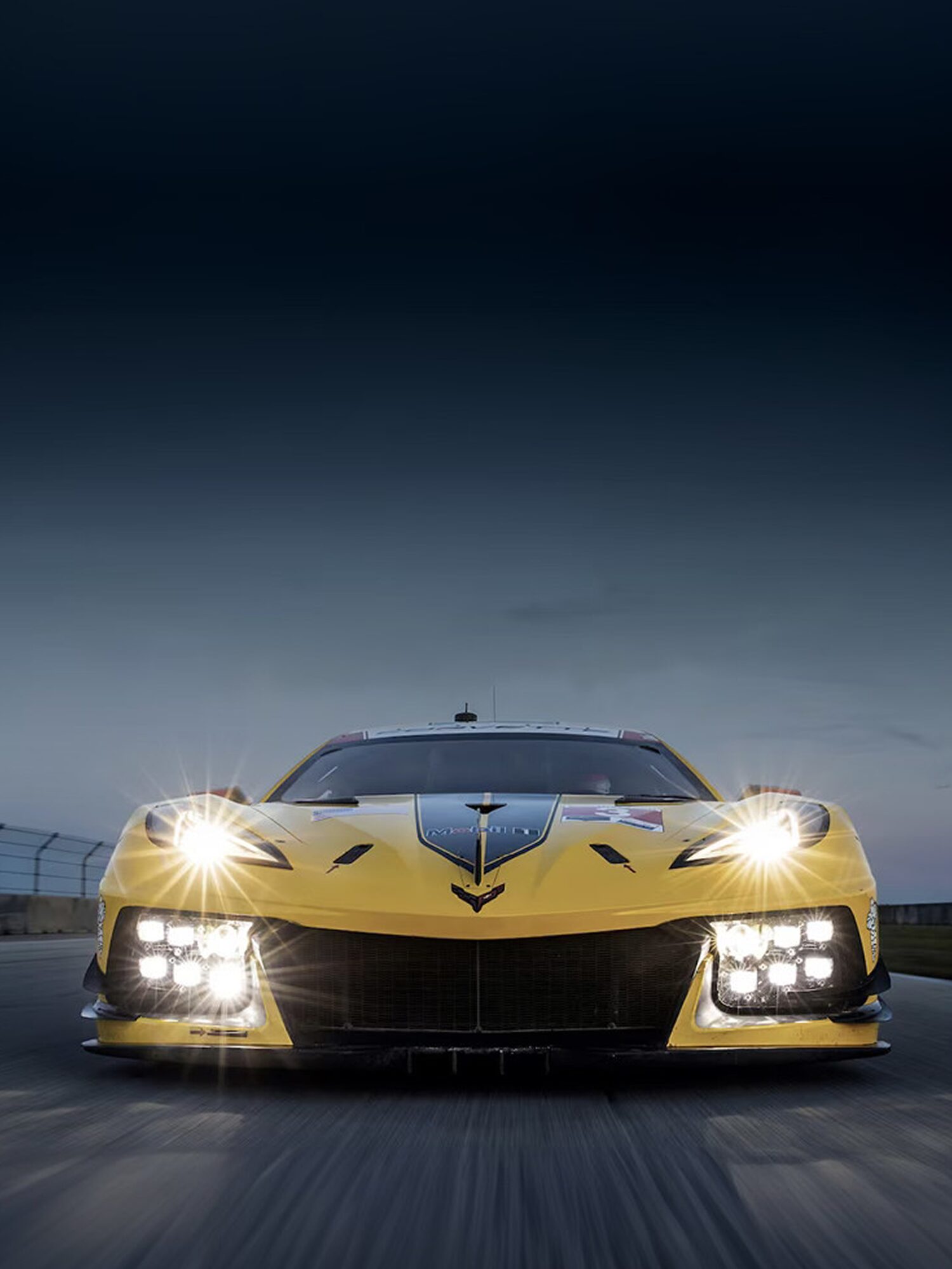A Yellow Chevy Corvette Race Car with Headlights On Driving on a Track at Dusk with Motion Blur.