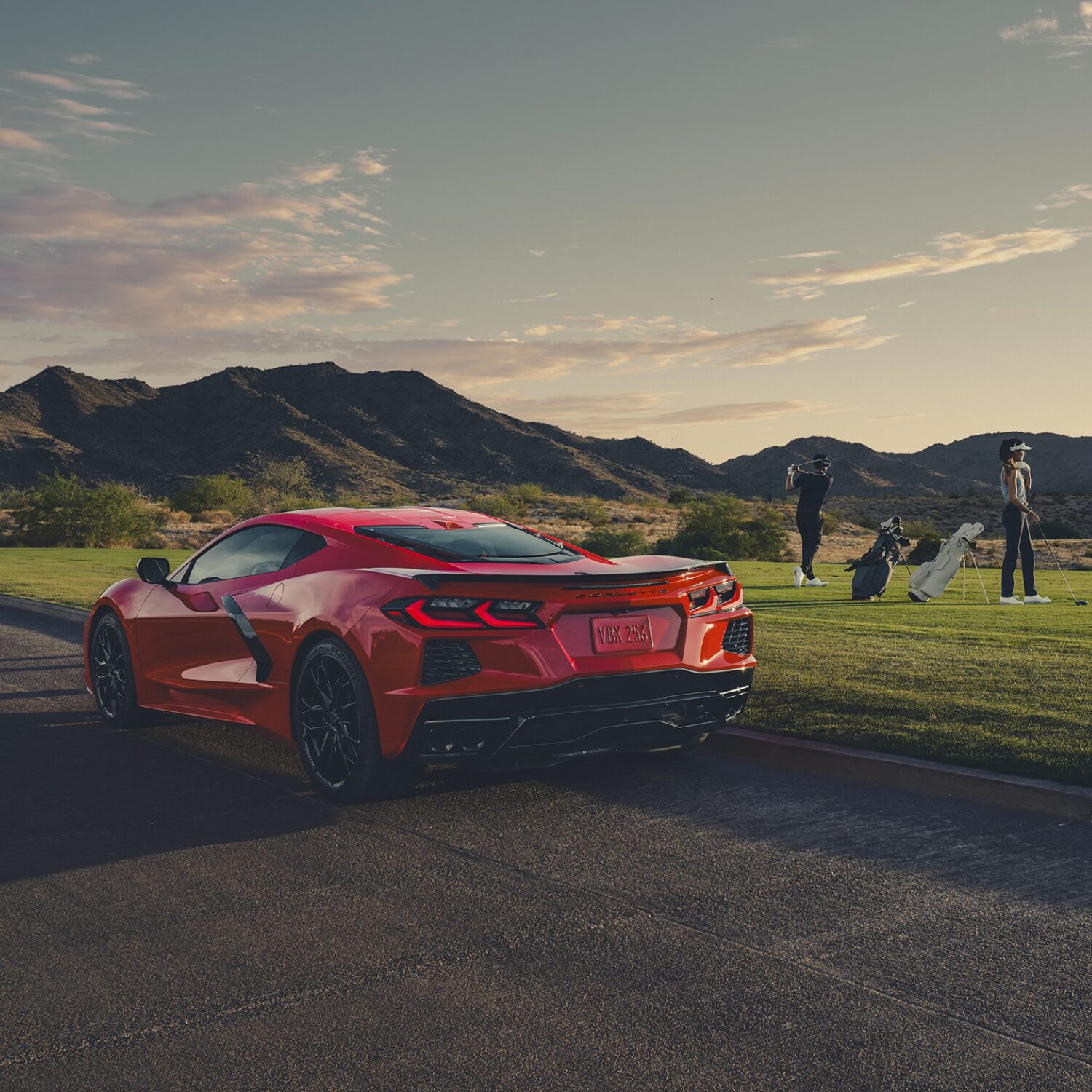 A Rear View of a Red Chevrolet Corvette Parked on a Paved Path Near a Golf Course with People Standing on the Grass and Mountains in the Background.