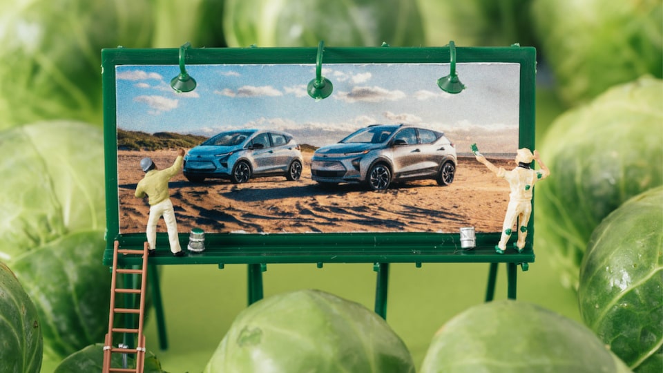 A diorama-like image of a miniature billboard showing the Bolt EV and Bolt EUV, with two tiny human figures painting it, surrounded by Brussels sprouts.