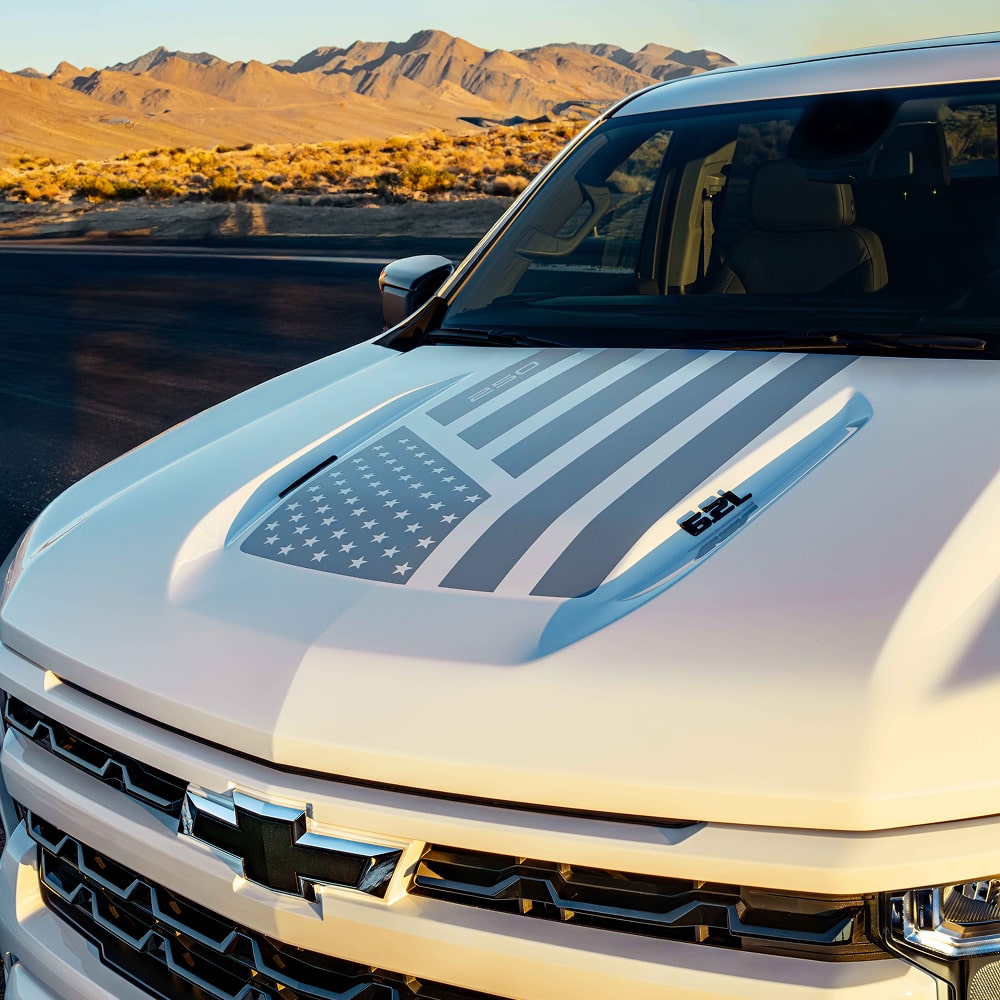 Front Angled View of a White Chevy Truck with a Gray Design of the American Flag on the Hood.