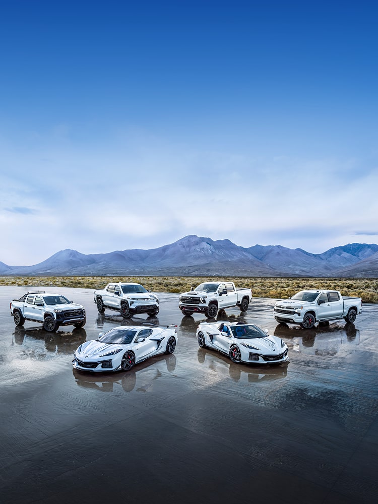 White Chevy Trucks and Corvettes Parked in a Field with Mountains in the Background.