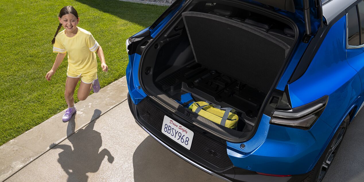 A Blue Chevrolet Equinox EV With Its Trunk Open, Revealing a Child in a Yellow Shirt Playing Nearby on a Driveway.