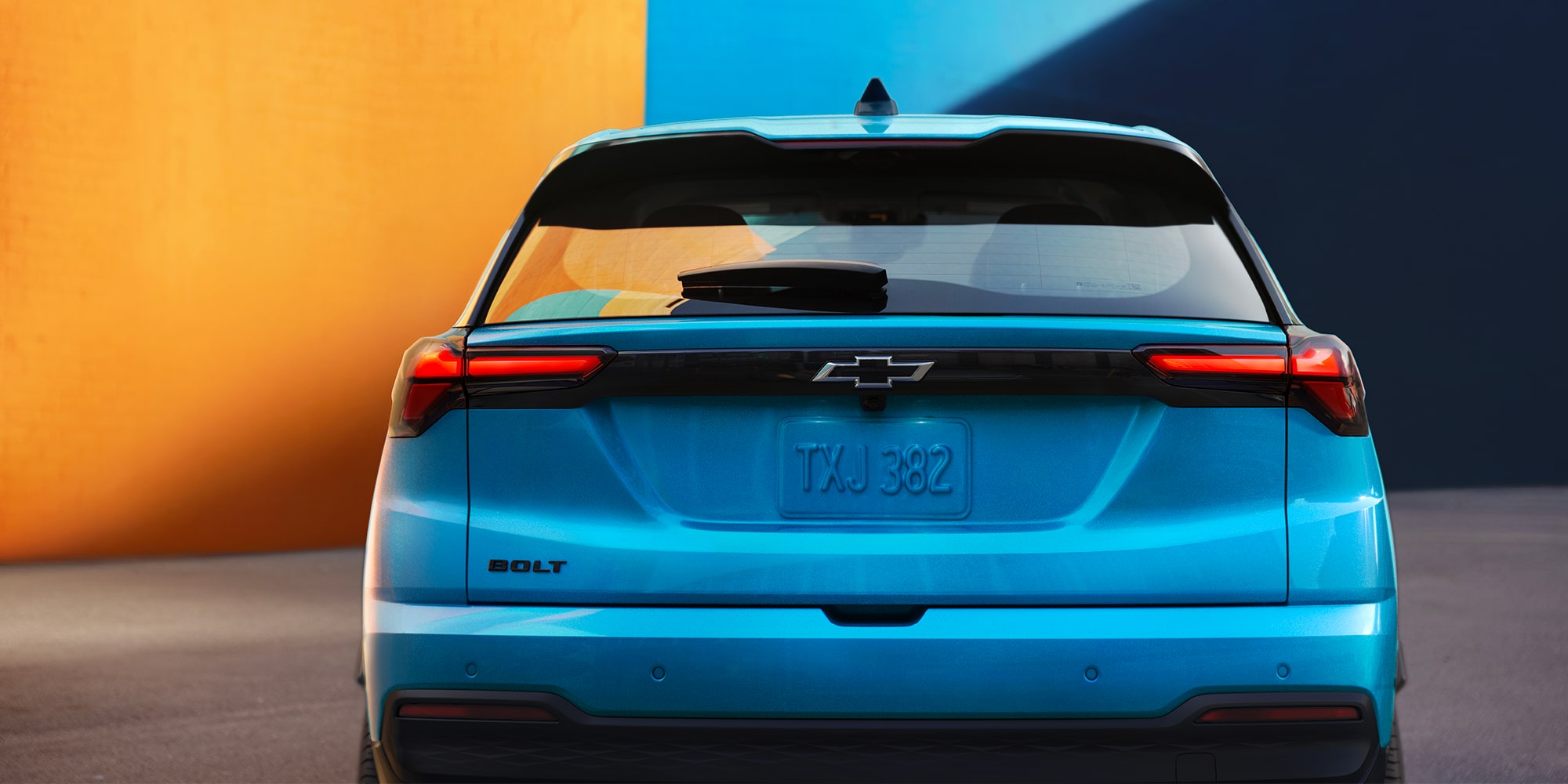 The Rear View of a Light Blue 2027 Chevrolet Bolt Shows the Vehicle Parked in Front of an Orange and Blue Wall, Highlighting the Updated Taillights and Design.