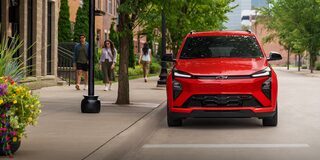 The Front of a Red 2027 Chevrolet Bolt is Shown in a City Setting, with People Walking on the Sidewalk and Buildings in the Background.