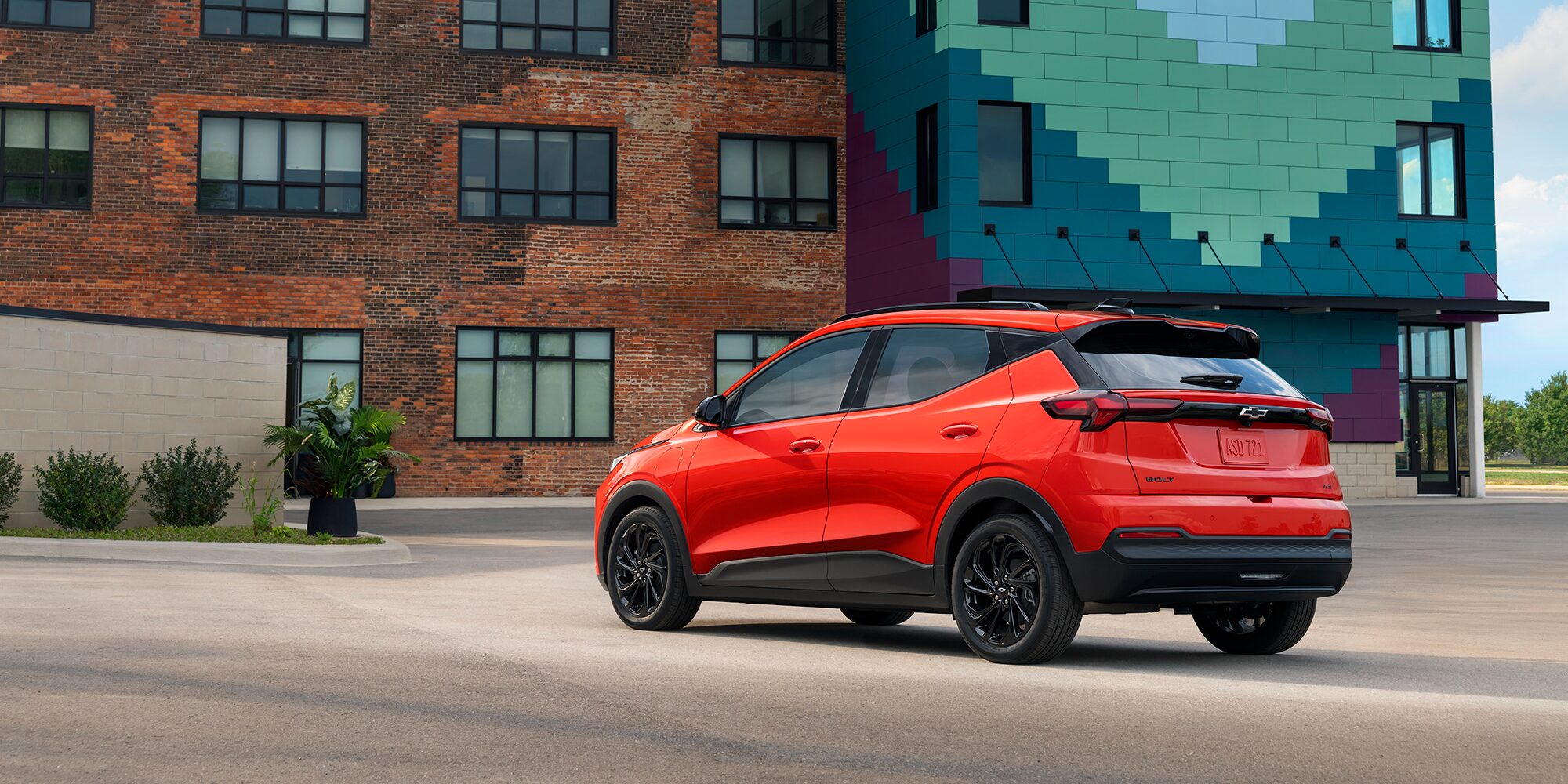 The Rear View of a Red 2027 Chevrolet Bolt is Shown in a Parking Lot, with a Modern Brick Building and a Multi-Colored Geometric-Patterned Building in the Background.
