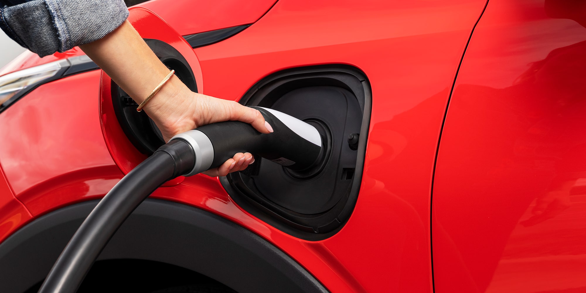 A Close-Up of a Hand Plugging a Charging Cable into the Charging Port of a Red 2027 Chevrolet Bolt.