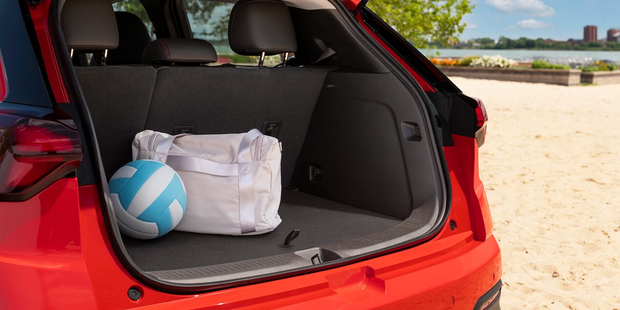 The Trunk of a Red 2027 Chevrolet Bolt is Open on a Sandy Beach, with a Blue and White Volleyball and a White Duffel Bag Inside.