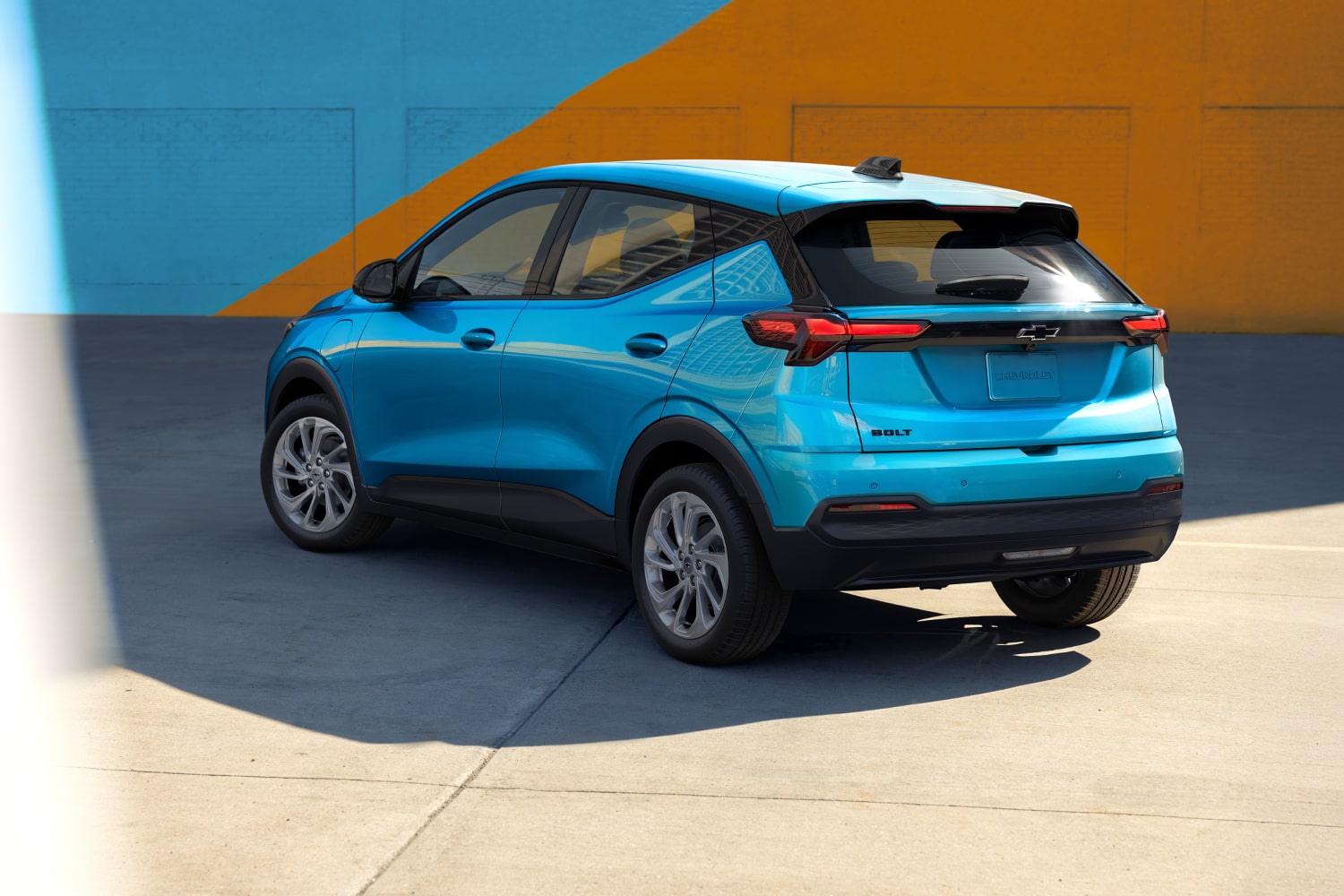 The Rear View of a Marina Blue Metallic 2027 Chevrolet Bolt Shows the Vehicle Parked in Front of a Blue and Orange Wall, Highlighting the Updated Taillights and Design.