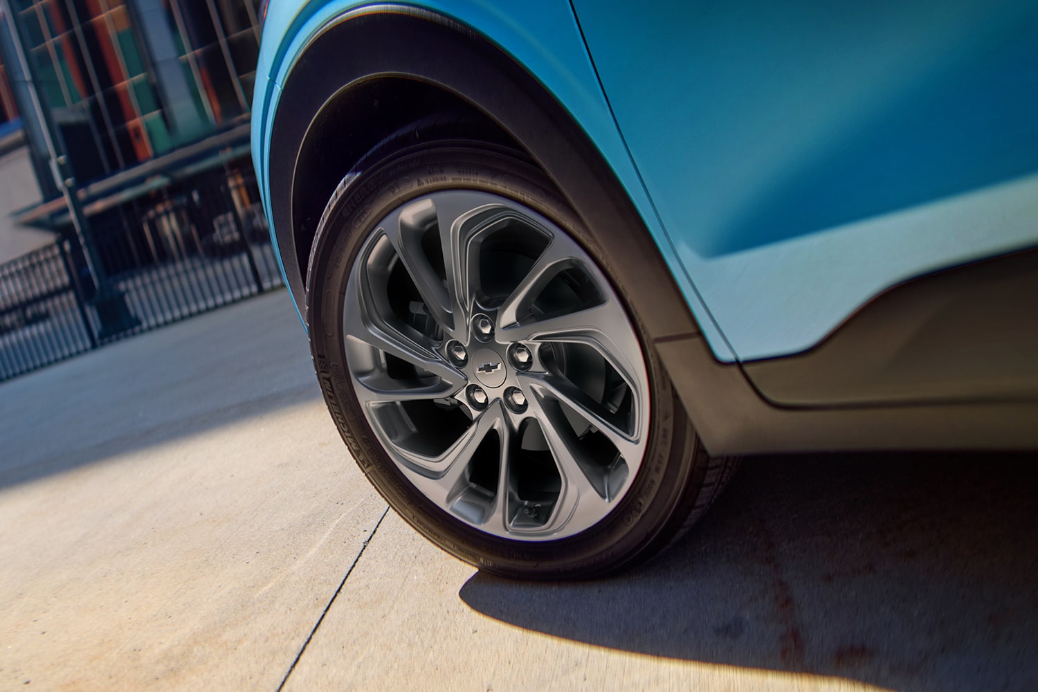 A Close-Up of the Front Wheel of a Marina Blue Metallic 2027 Chevrolet Bolt, Showing the Multi-Spoke Aluminum Wheel with a Chrome Chevrolet Bowtie in the Center.