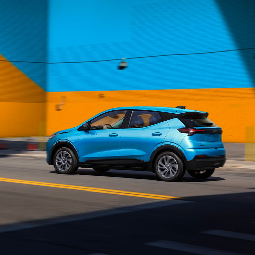 The Rear View of a Marina Blue Metallic 2027 Chevrolet Bolt Shows the Vehicle Parked in Front of a Blue and Orange Wall, Highlighting the Updated Taillights and Design.