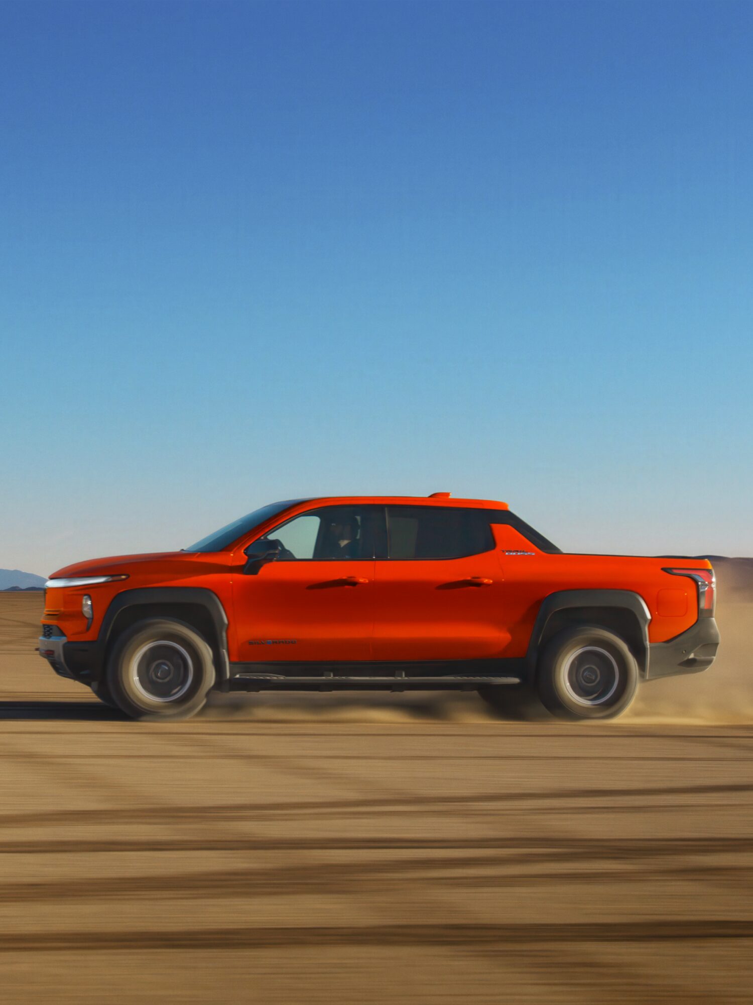 Side View of Orange 2026 Silverado EV Driving on a Dirt Road Highlighting its Off-Road Capabilities and Sleek Design