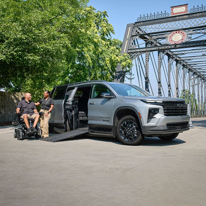 A Silver Chevy Traverse SUV With an Extended Wheelchair Ramp Parked on a Road Beside a Steel Bridge While Two People Converse Near the Ramp.