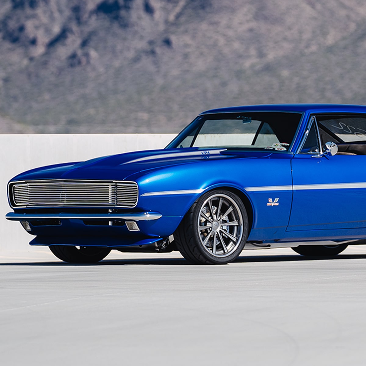 A Blue Classic Chevrolet Camaro SS with Chrome Accents and Polished Wheels Parked on a Light Surface in Front of a Mountain Landscape.