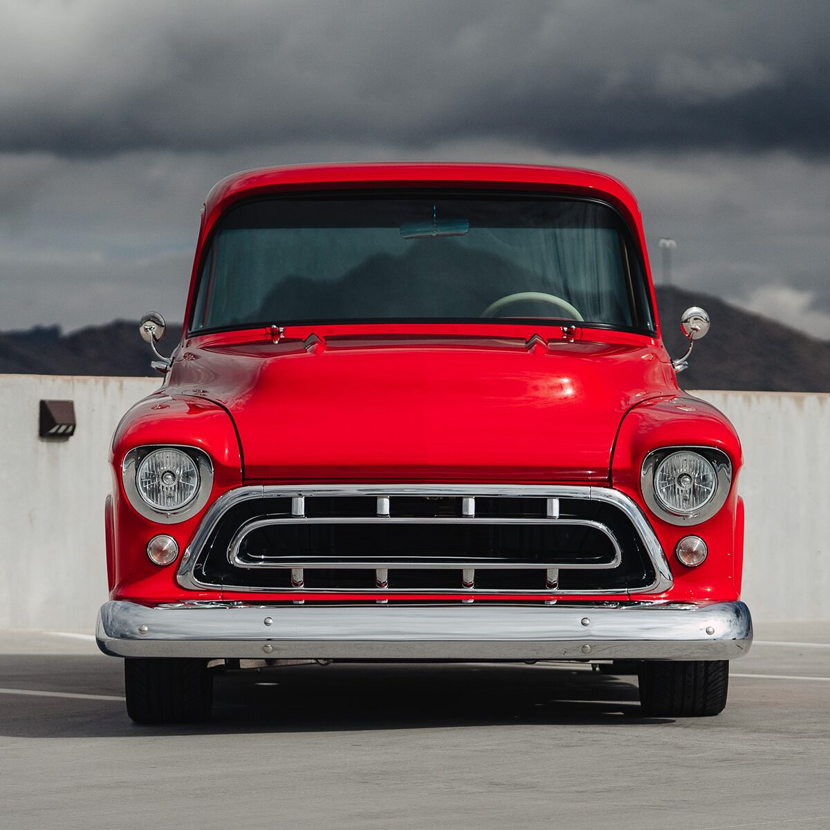 A Front View of a Red Chevrolet 3100 Pickup Truck Parked in Front of a Half Cement Wall and a Grey Sky with Mountains behind it. 