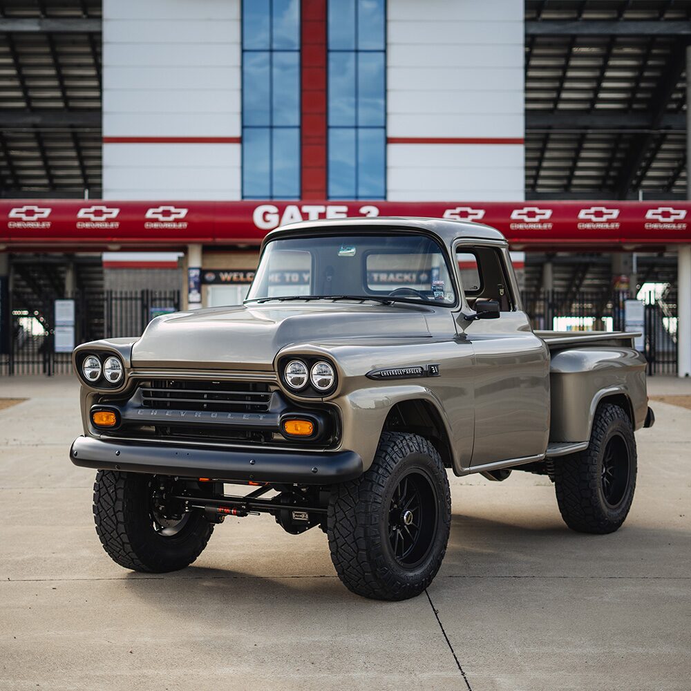 A Side Angled View of a Silver 1958 Chevrolet Apache Parked in Front of the Entry Way of a Race Track.
