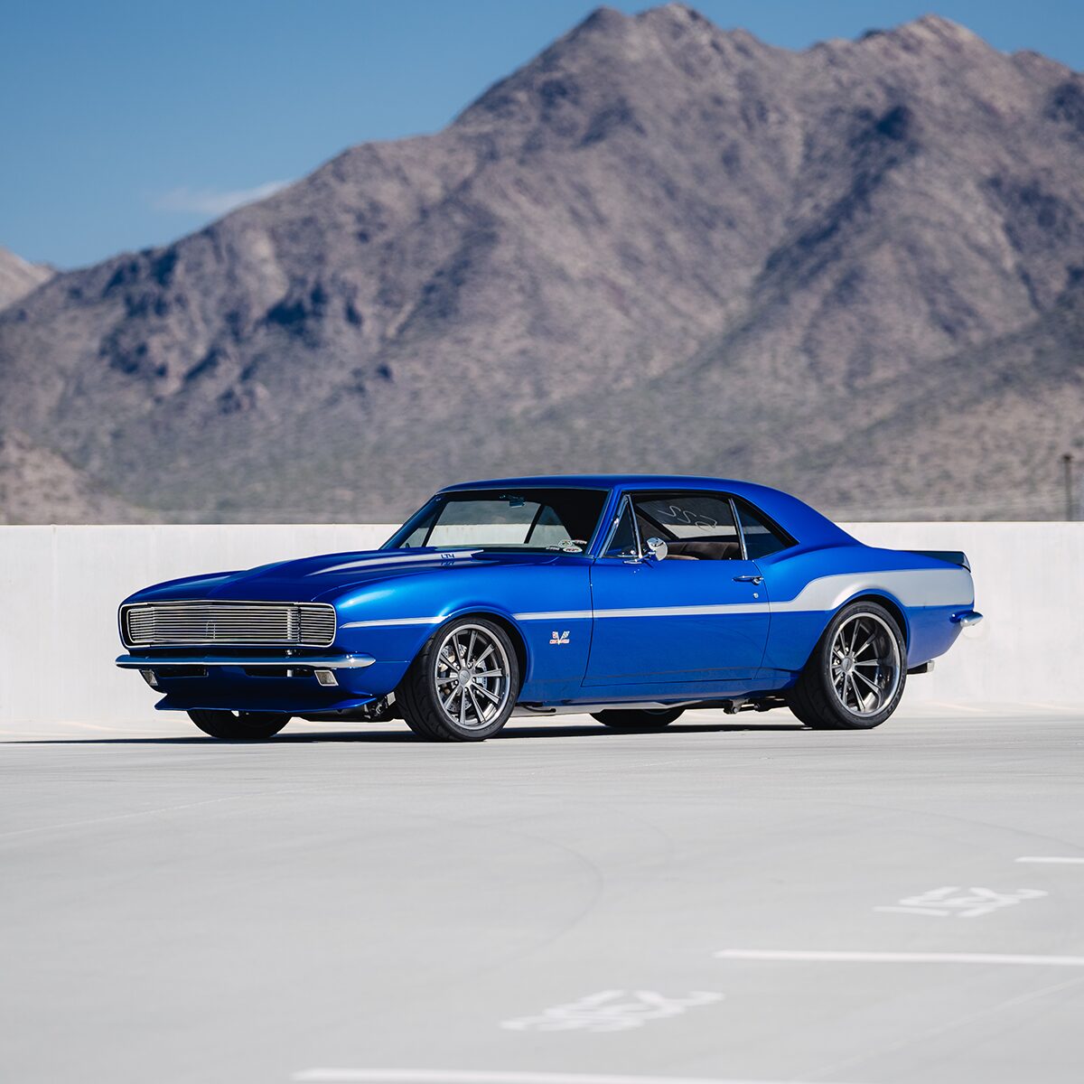 An Exterior Profile View of a Blue 1967 Chevrolet Camaro SS Parked in a Parking Lot with Mountains in the Background.