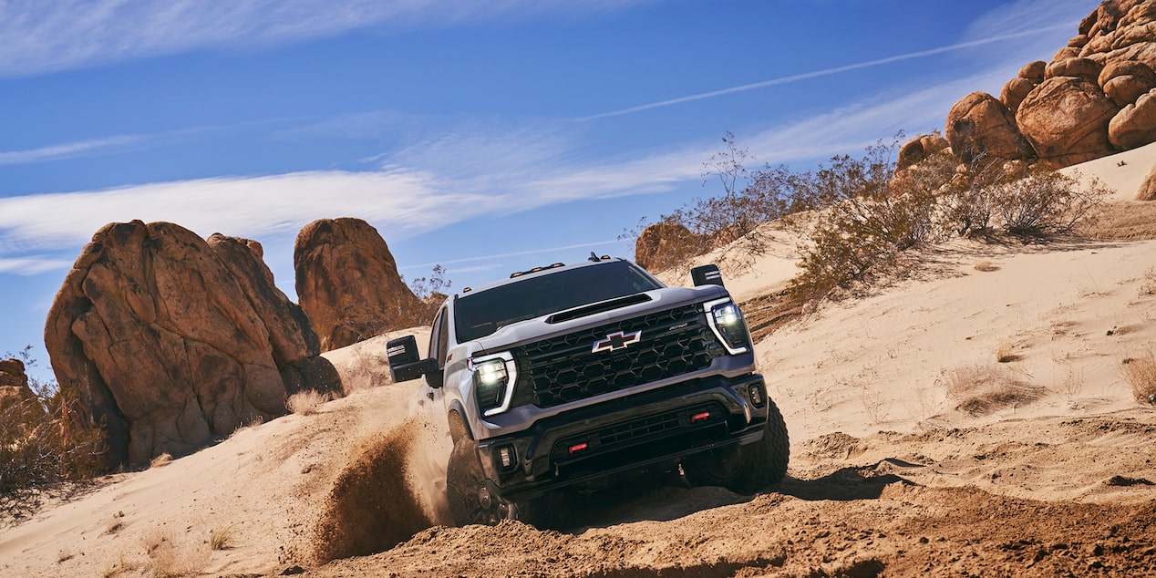 A Black Chevy Silverado LD Climbing Over Large Desert Rocks with Dust and Sand Kicking Up from Its Tires.