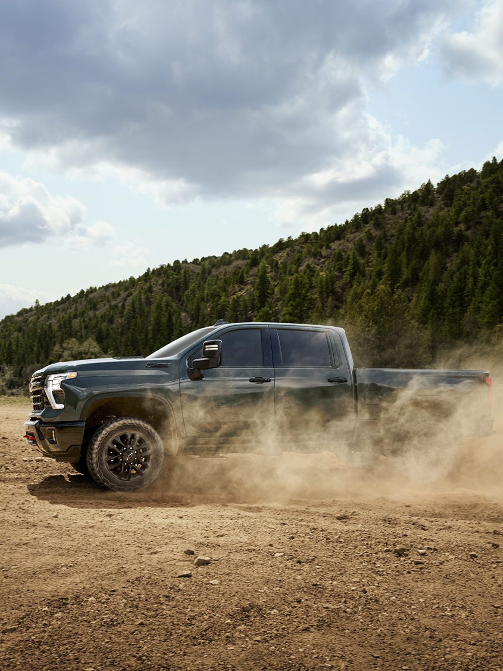 A Blue Chevy Silverado LD Driving Through a Dusty Dirt Road Surrounded by Tall Green Pine Trees and Mountains in the Distance.