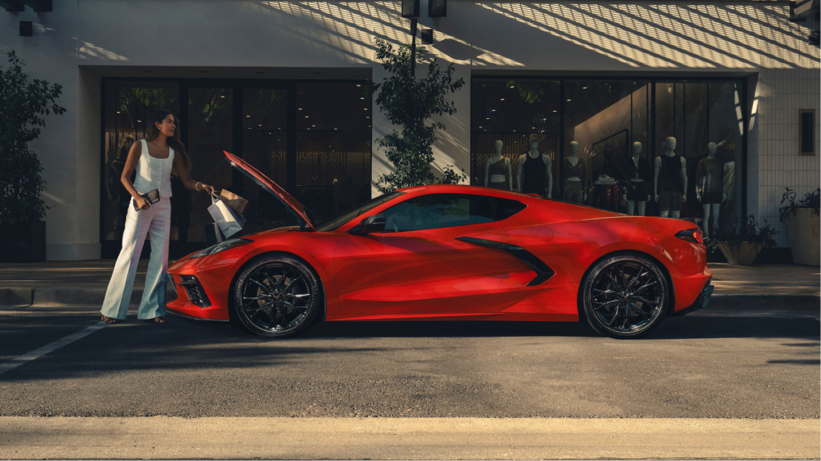 Side Profile of a 2025 Corvette Stingray Parked Outside a Store Front and Woman Placing Bags in Front Trunk