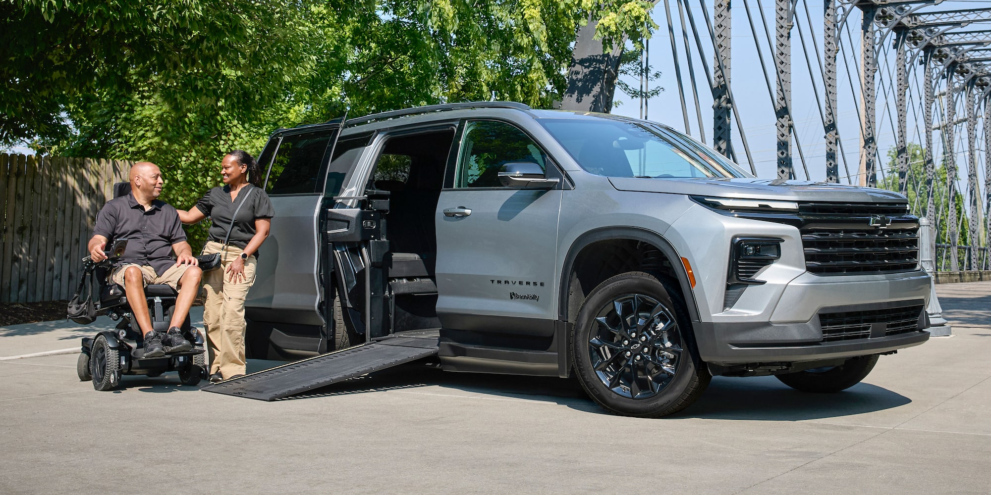 A Silver Chevy Traverse SUV With an Extended Wheelchair Ramp Parked on a Road Beside a Steel Bridge While Two People Converse Near the Ramp.