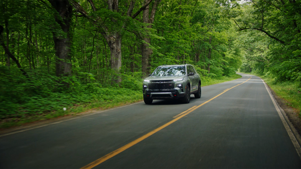 Far Away Front View of a 2025 Chevy Traverse Driving Down the Road Surrounded by Trees
