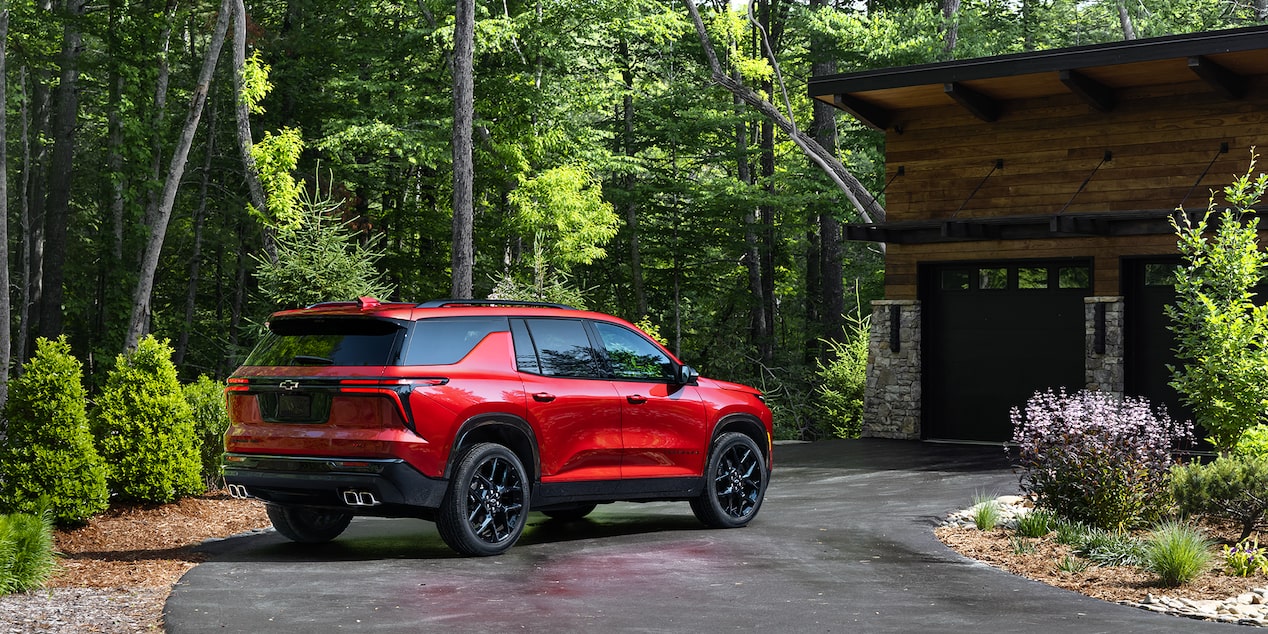 Three Quarters View of a Red 2025 Chevy Traverse Infront of a Wooden House Surrounded by Trees