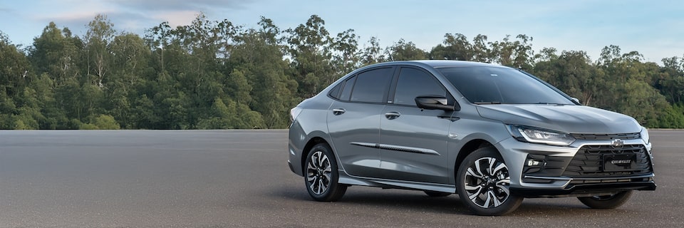 Chevrolet Onix Plus em vista de frente e lateral direita estacionado em pista aberta com vegetação ao fundo.