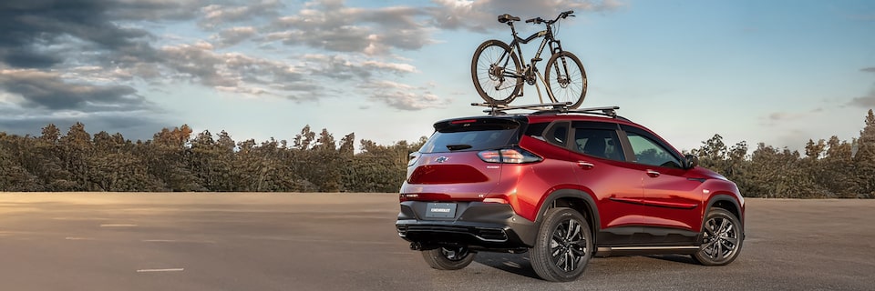 Chevrolet Tracker em vista de traseira e lateral direita com bicicleta montada no teto em pista com vegetação ao fundo.