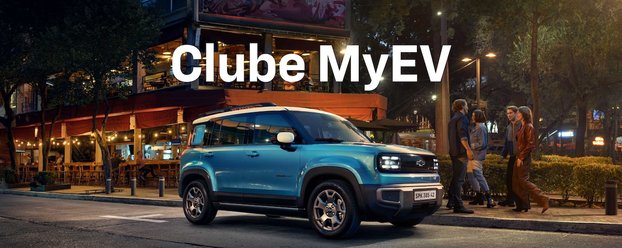 SUV Chevrolet elétrico azul estacionado à noite em frente a um restaurante iluminado, com grupo de pessoas conversando ao fundo e texto Clube MyEV.