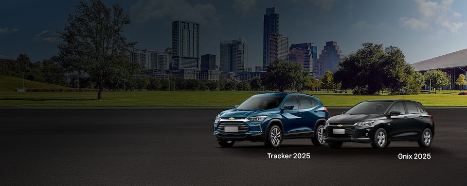 Dois veículos Chevrolet, um Onix preto e um Tracker azul, estão expostos em uma rua larga. Prédios urbanos modernos e árvores verdes estão ao fundo.