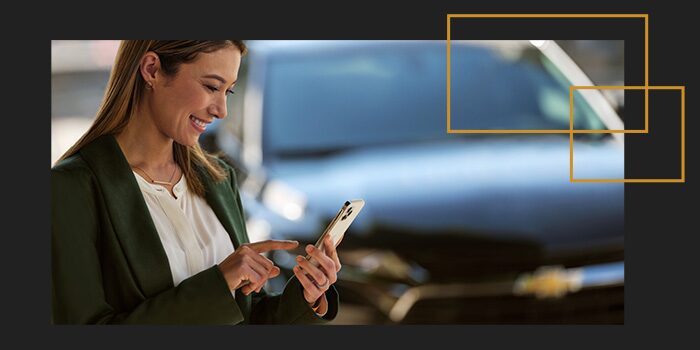 Mulher branca, de cabelos claros, vestida como executiva, de perfil, na calçada, olhando o celular e sorrindo. Ao fundo, um Chevrolet estacionado.