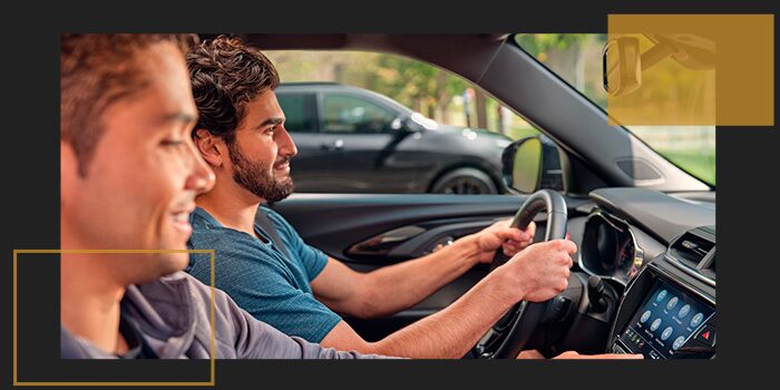 Dois homens brancos, sorrindo, um dirigindo e o outro no banco do passageiro, acionando o central multimídia, à bordo de um Chevrolet.