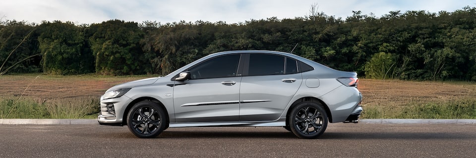 Chevrolet Onix Plus em vista lateral esquerda exibido em pista com vegetação ao fundo.