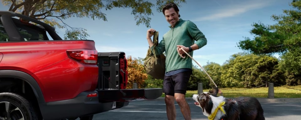 Um homem com roupas de treino e segurando um cachorro pela coleira está ao lado de uma Chevrolet Montana vermelha enquanto coloca sua mochila na caçamba. Ao fundo, temos arbustos e árvores, dando a entender que se trata de um parque.