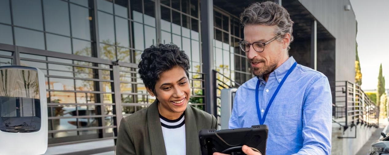 vemos duas pessoas em frente à uma concessionária Chevrolet. Um vendedor, usando óculos, camisa e crachá azul está mostrandos os modelos em um tablet para uma mulher, que usa uma camiseta branca e um blazer cinza