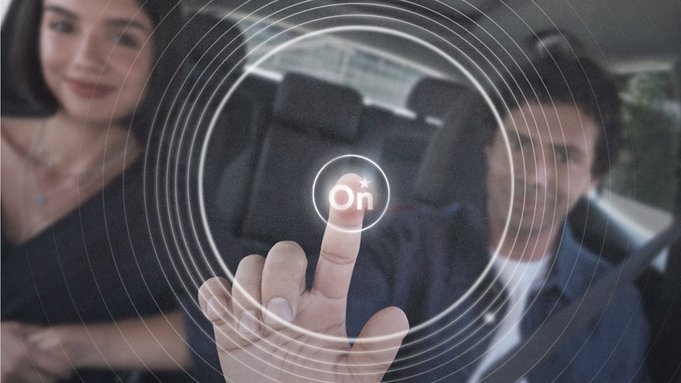 Homem branco no banco do motorista tocando virtualmente um botão “On” iluminado no ar, com efeito gráfico circular ao redor; uma mulher branca está sentada ao seu lado no banco do passageiro.