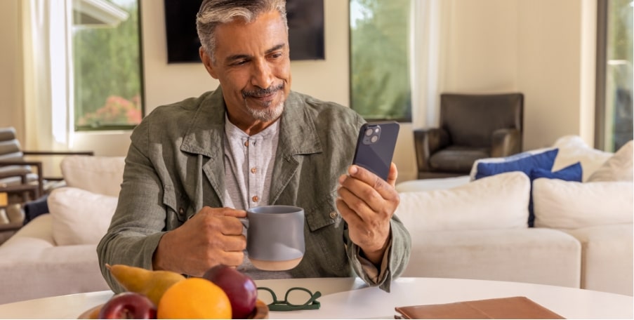 Imagem de um homem sentado à mesa, segurando uma xícara com uma mão e usando o celular com a outra, acessando o OnStar.