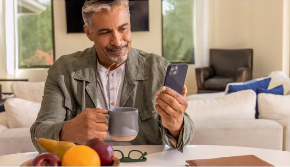 Imagem de um homem sentado à mesa, segurando uma xícara com uma mão e usando o celular com a outra, acessando o OnStar.