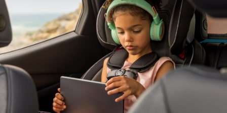 Imagem de uma menina negra sentada no banco de trás do carro , com fone de ouvindo mexendo no tablet.