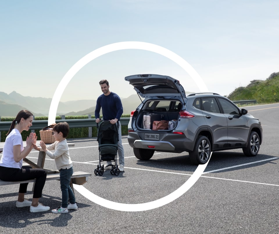 Imagem de uma família ao ar livre, brincando ao lado de um veículo Chevrolet, que está com o porta-malas aberto.