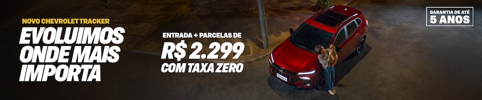 Novo Chevrolet Tracker, cor vermelha, visto de cima em uma rua à noite. Uma mulher segura um cão pequeno ao lado do carro. Texto promocional: Evoluímos onde mais importa. Taxa zero e parcelas de apenas R$ 2.299 com 5 anos de garantia. Selo Garantia de até 5 anos no canto inferior direito.