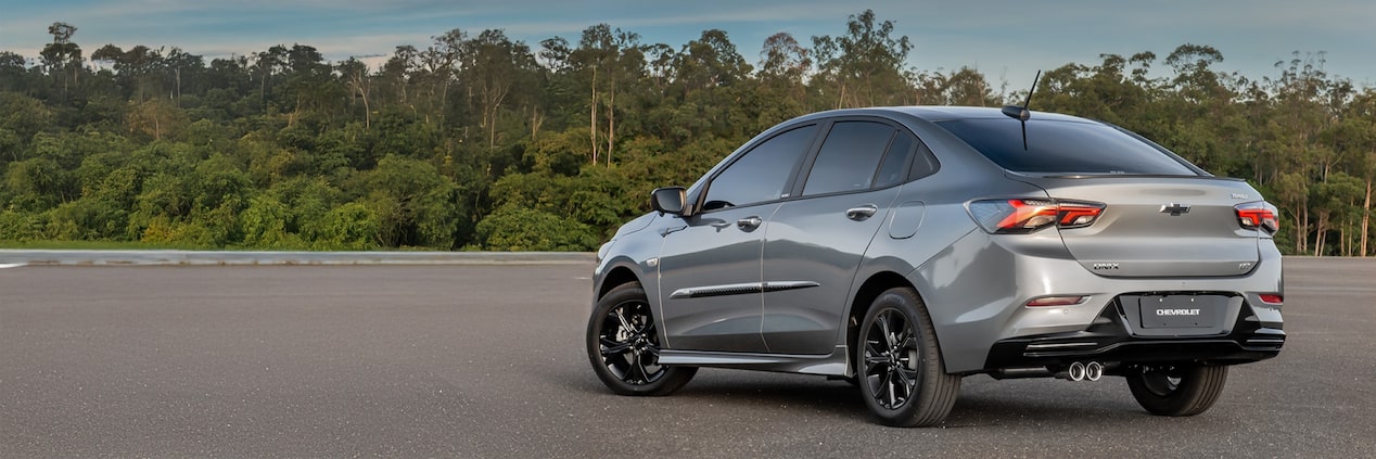 Chevrolet Onix Plus em fotografia embientada. O carro, na cor prata e em posição 3/4 de costas, está estacionado diante de uma parede de árvores altas e bem verdes. No quadrante superior esquerdo, o texto: Onix - Acessórios.