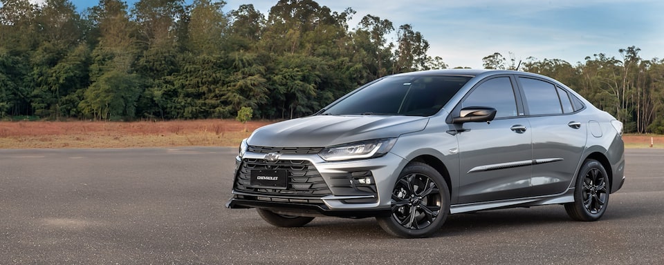 Chevrolet Onix Plus em fotografia embientada. O carro, na cor prata, está estacionado diante de uma parede de árvores altas e bem verdes. No quadrante superior esquerdo, o texto: Onix - Acessórios.