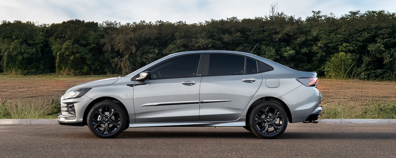 Chevrolet Onix Plus na cor prata em fotografia ambientada. O veículo é visto lateralmente e ilustra a página de acessórios originais Chevrolet dedicada a ele próprio. No quadrante superior esquerdo, o texto: Onix - Aparência.