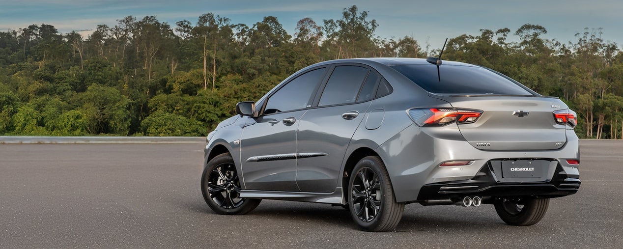 Masthead versão desktop da página de Pacotes, dentro de acessórios originais Chevrolet, para o Onix Plus, que aparece em foto ambientada, estacionado diante de uma grande parede de árvores verdinhas.