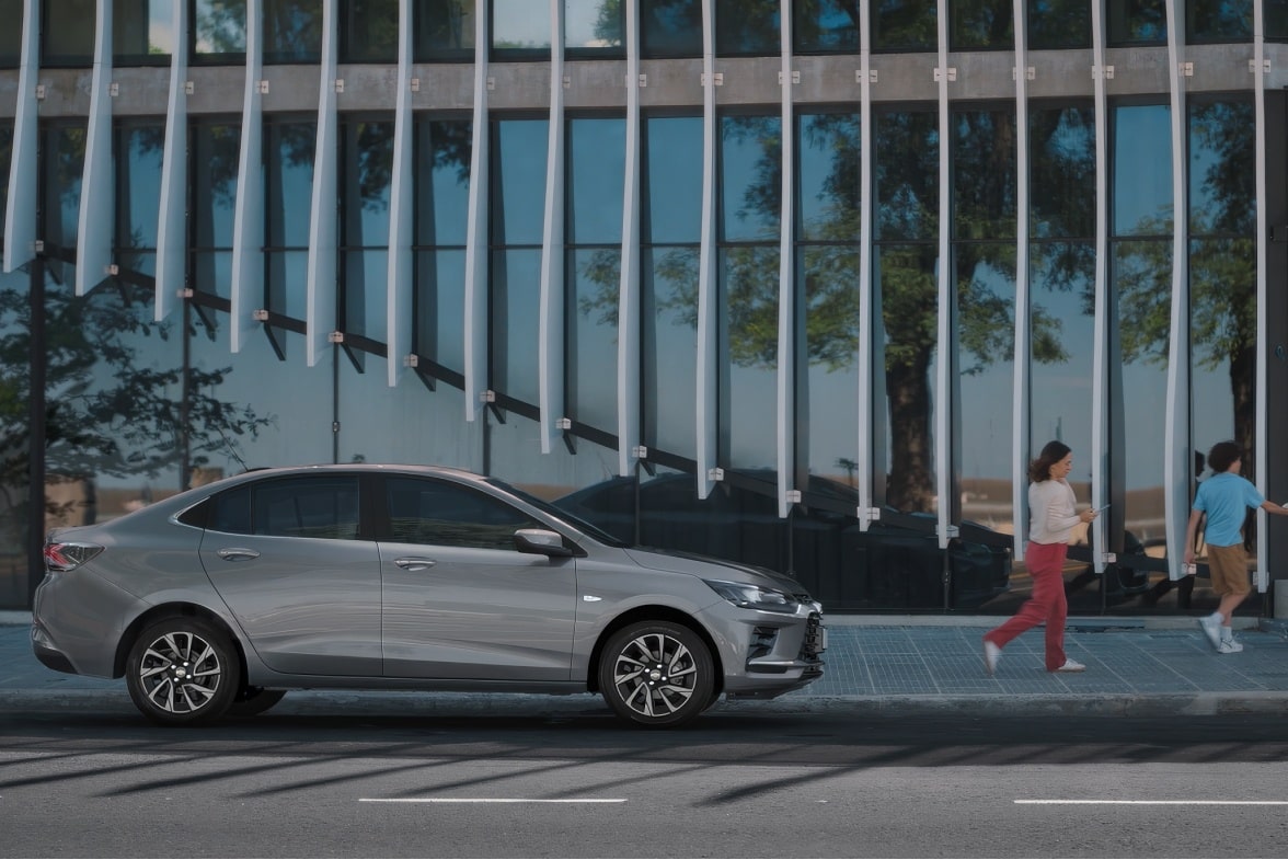Chevrolet Onix Plus na cor Prata Shark estacionado em frente a fachada moderna, com duas pessoas caminhando ao lado.