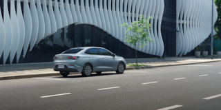 Chevrolet Onix Plus na cor Prata Shark circulando em avenida com fachada arquitetônica de linhas onduladas ao fundo.