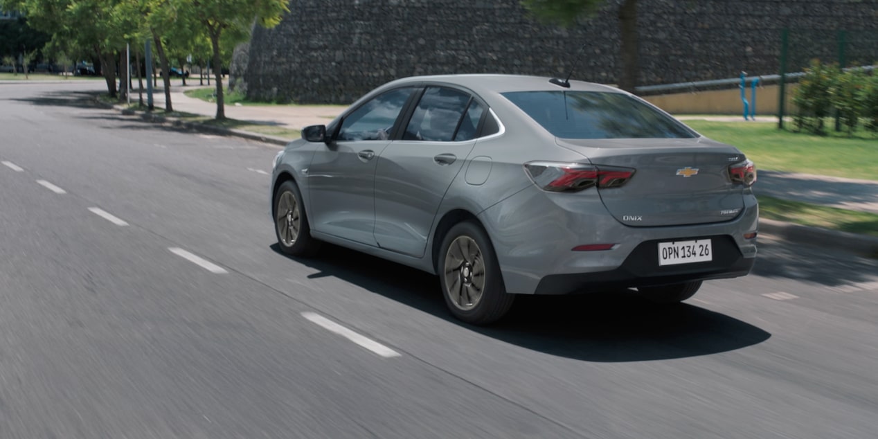 Vista traseira do Chevrolet Onix Plus na cor Prata Shark, em movimento com muro de pedra ao fundo.