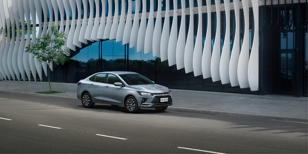Chevrolet Onix Plus na cor Prata Shark em avenida com fachada arquitetônica de linhas onduladas ao fundo.